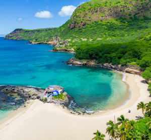 Bucht mit wei&szlig;em Sandstrand, t&uuml;rkisfarbenem Wasser und gr&uuml;n bewachsenen H&uuml;geln auf den Kapverdischen Inseln unter blauem Himmel.