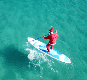 Als Weihnachtsmann verkleideter Mann paddelt auf einem Surfbrett im türkisblauen Meer in Australien.