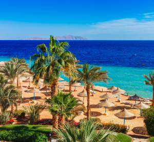 Strandresort in Hurghada mit Palmen, Sonnenschirmen aus Stroh und türkisblauem Meer unter klarem Himmel.