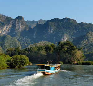 Langsames Holzboot f&auml;hrt auf einem breiten Fluss, umgeben von dicht bewaldeten Bergen im goldenen Abendlicht.