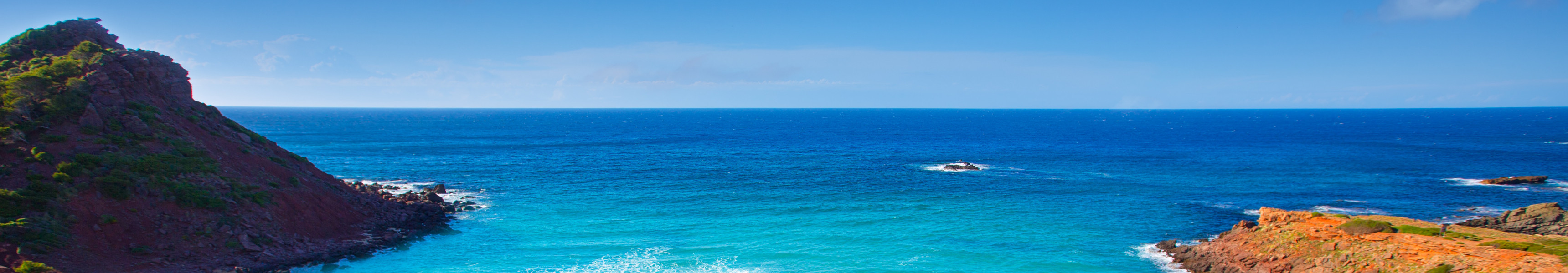 Unberührter Strand Cala Pregonda auf Menorca mit goldenem Sand, türkisblauem Meer und rötlichen Felsen unter blauem Himmel
