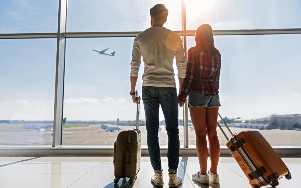 Reisepaar am Flughafen hält Händchen und blickt durch die große Glasfassade auf ein startendes Flugzeug, bereit für ihr nächstes Abenteuer.