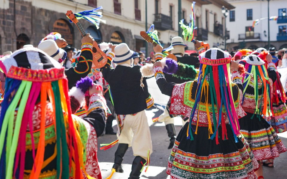 Teilnehmende des Inti Raymi-Festes in traditioneller Kleidung tanzen und musizieren auf einer engen Stra&szlig;e, geschm&uuml;ckt mit bunten B&auml;ndern und Instrumenten, w&auml;hrend Zuschauer das Spektakel verfolgen.