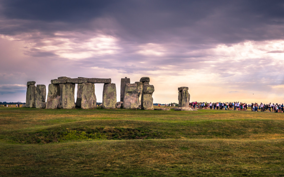 Panoramablick auf Stonehenge unter einem dramatischen, bew&ouml;lkten Himmel, eine Gruppe von Besuchern betrachtet das historische Monument aus einiger Entfernung.