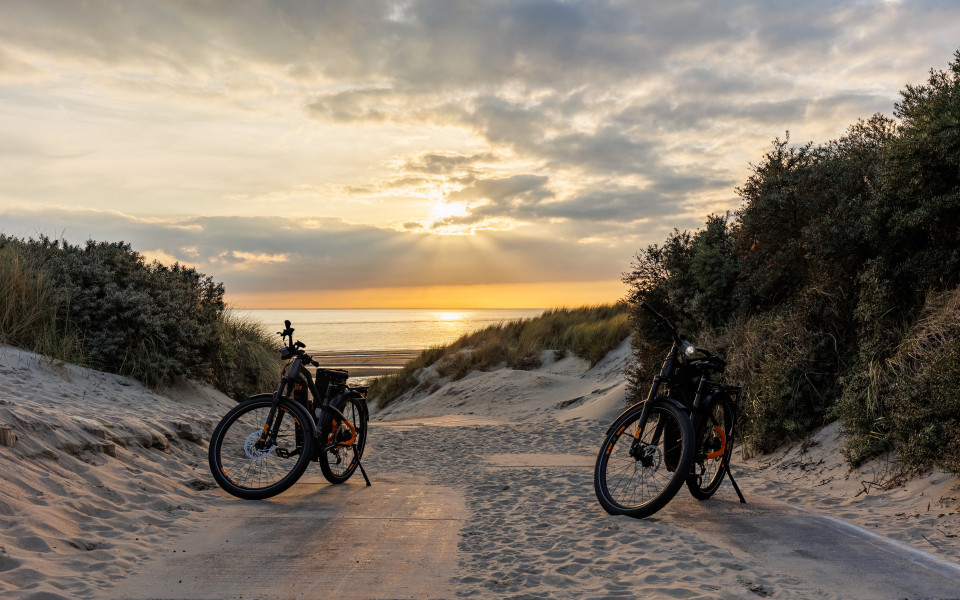 Zwei Fahrräder stehen in einer Dünenlandschaft auf einem sandigen Weg mit Blick auf das Meer und einen Sonnenuntergang am Horizont.