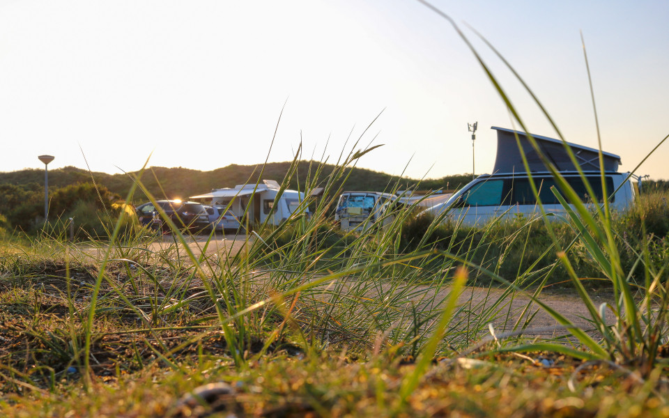 Gras wächst neben einer Straße, die durch einen Campingplatz in den Dünen Nordhollands führt. Im Hintergrund stehen Wohnmobile, Wohnwagen und Autos auf den Campingplätzen