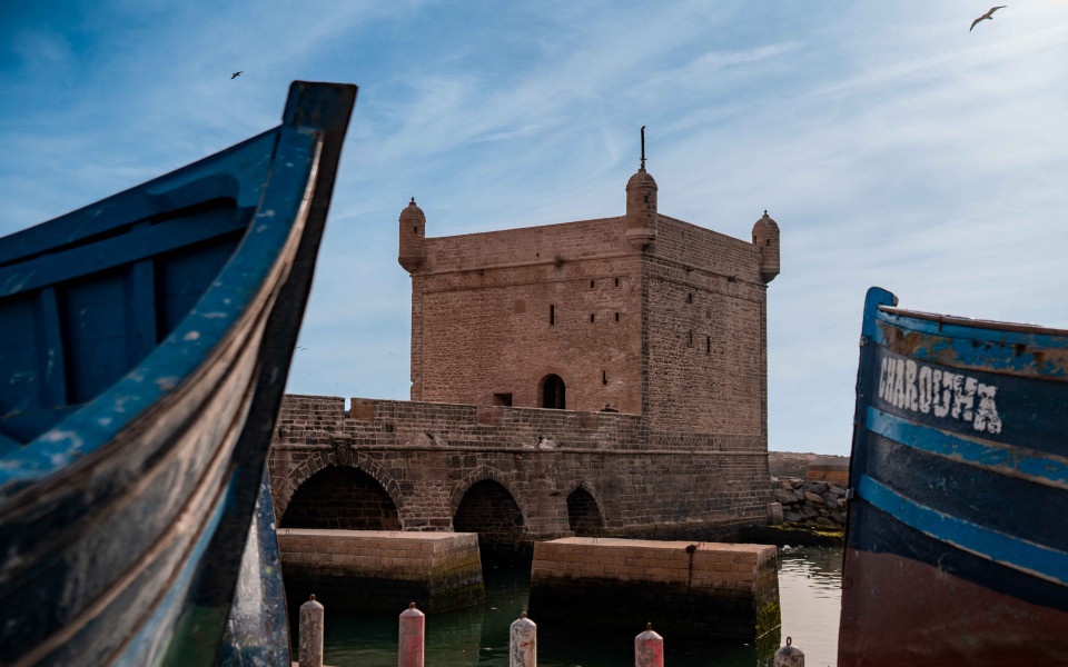 Blick auf das historische Stadttor Skala de la Ville in Essaouira, eingerahmt von zwei traditionellen blauen Fischerbooten im Hafen. 
