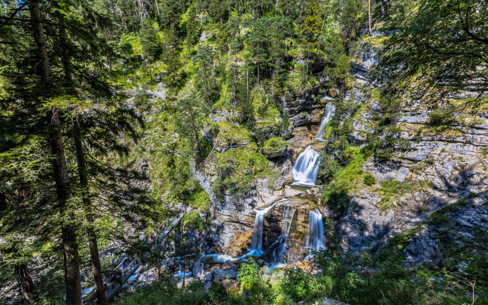 Mehrstufiger Wasserfall inmitten eines dichten Waldes mit Holzstegen und Wanderwegen, eingebettet in eine felsige Schlucht bei strahlendem Sonnenschein. 