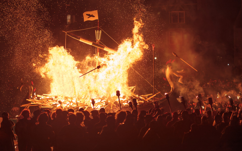 Ein gro&szlig;es, brennendes Wikingerschiff bei einem Feuerfest in der Nacht, mit vielen Zuschauern und Fackeln.