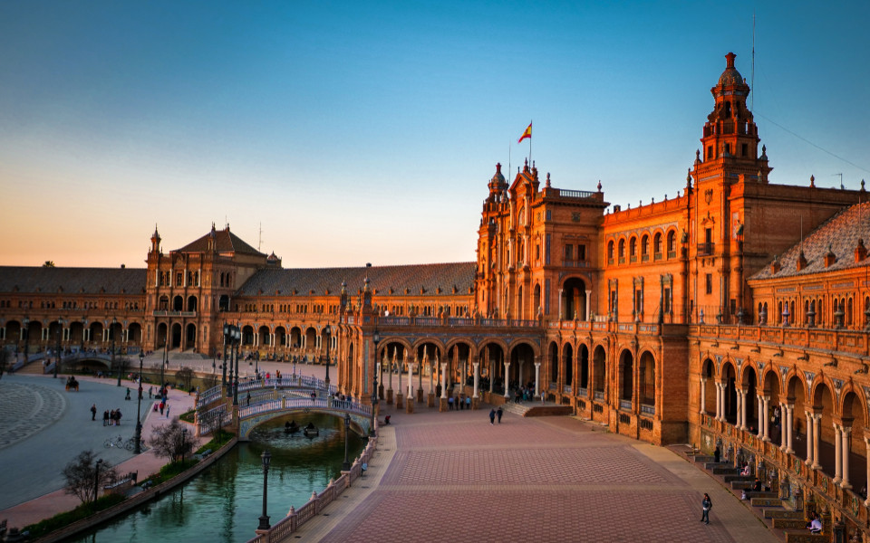 Panoramablick auf die Plaza de Espa&ntilde;a in Sevilla bei warmem Abendlicht, mit halbrundem Backsteingeb&auml;ude, Arkadeng&auml;ngen, Br&uuml;cken &uuml;ber einen Kanal und blauem Himmel.
