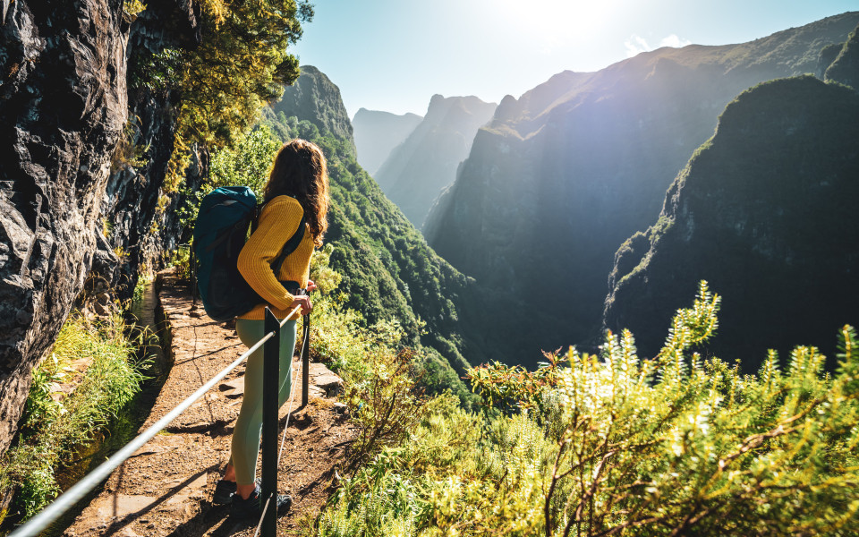Eine Rucksacktouristin genie&szlig;t die malerische Aussicht von unterhalb einer gro&szlig;en Felswand entlang eines Wasserlaufs an einer steilen Klippe im Regenwald von Madeira. 